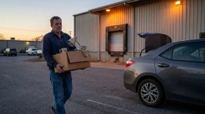 man carrying cardboard box with desk lamp toward open car trunk outside warehouse
